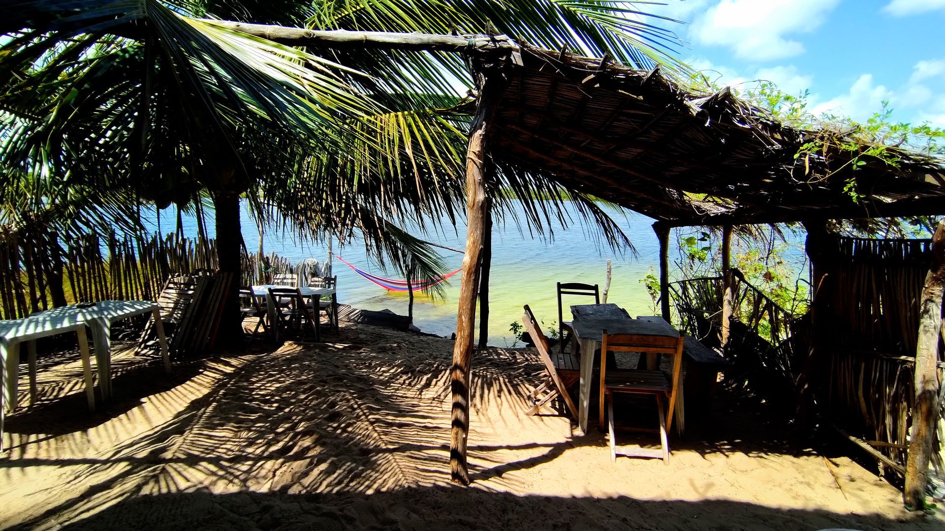 Uma cabana de praia rústica, com telhado de palha, mesas e cadeiras, com vista para um oceano ensolarado e turquesa.
