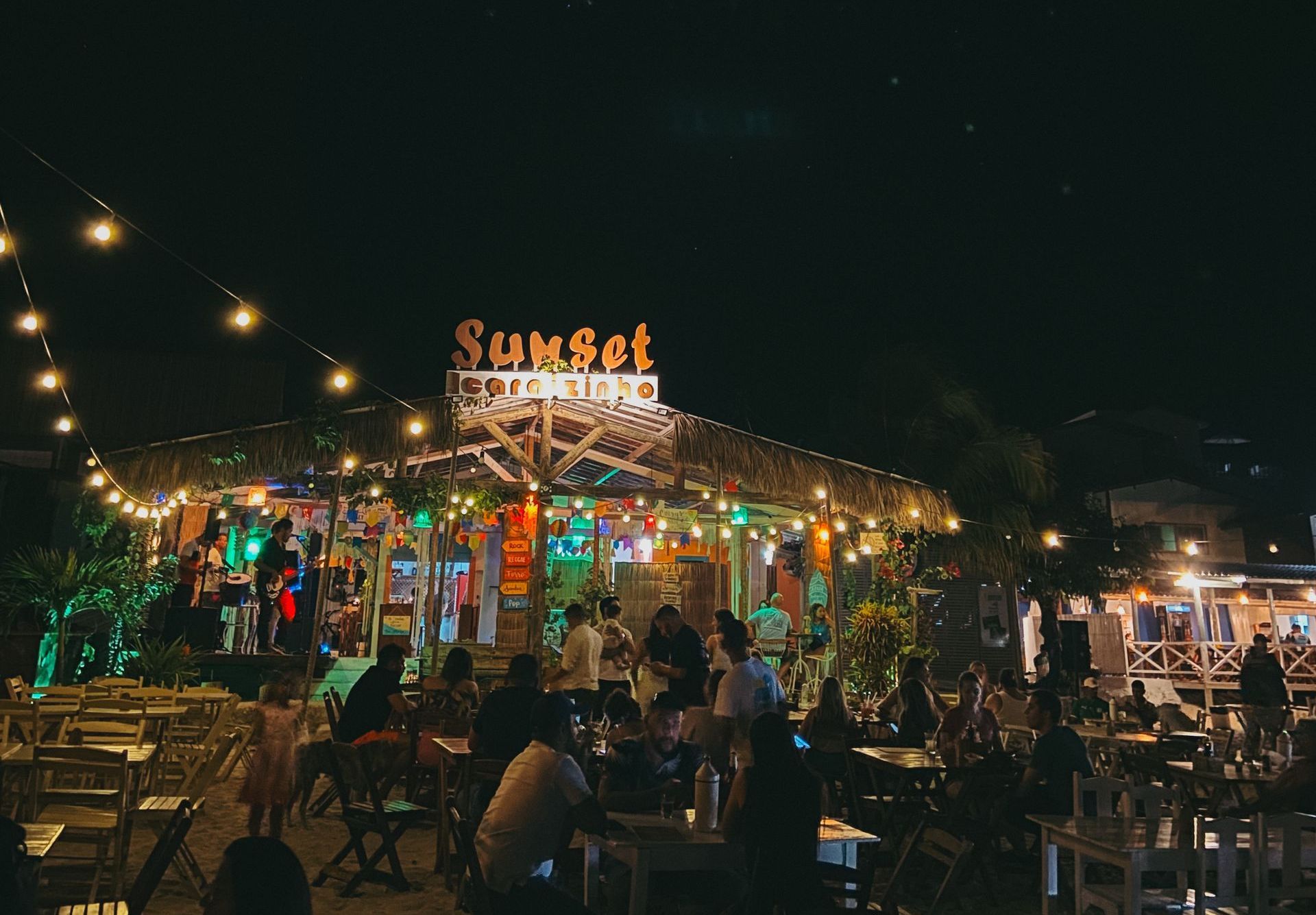 Restaurante ao ar livre à noite com luzes de corda aconchegantes, uma placa iluminada com a palavra 