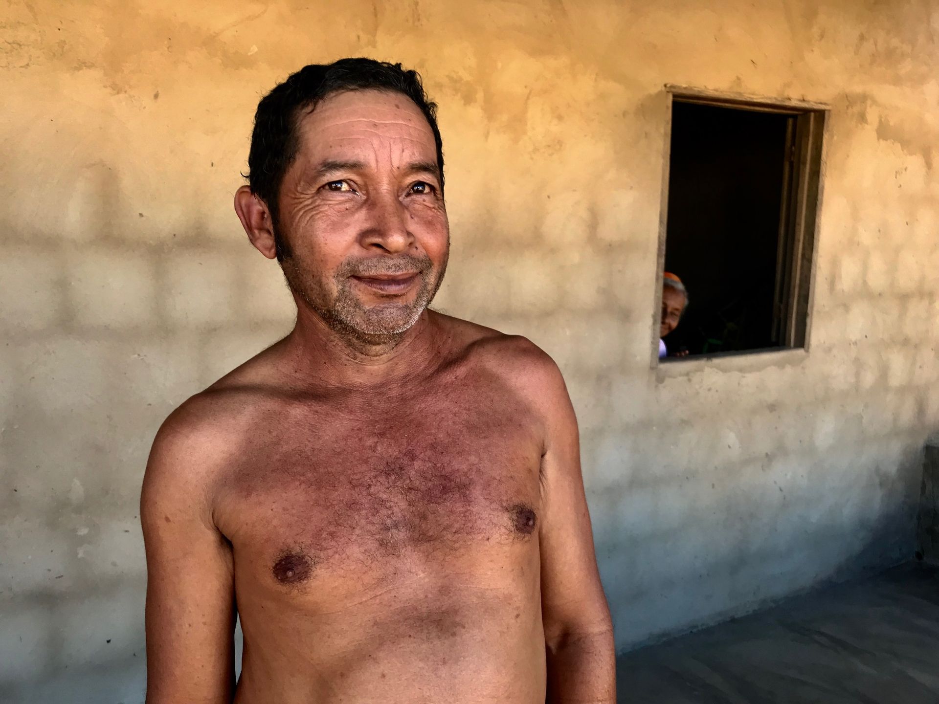 Uma pessoa com cabelo curto e escuro e barba rala, sem camisa, está de pé em frente a uma parede bege com uma janela escura ao fundo.