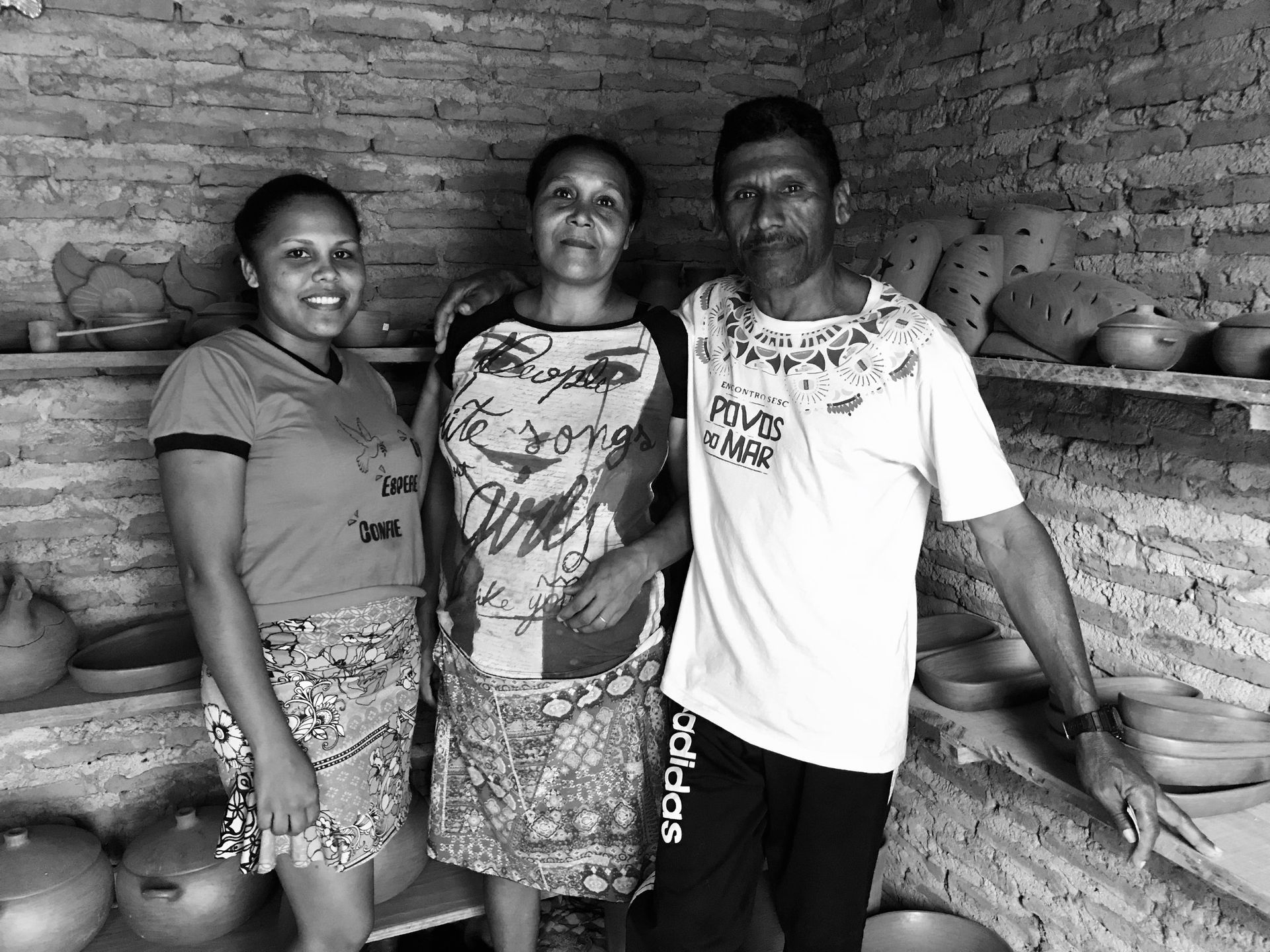 Três pessoas estão sorrindo em uma sala com paredes de tijolos, repleta de peças de cerâmica artesanal em prateleiras.