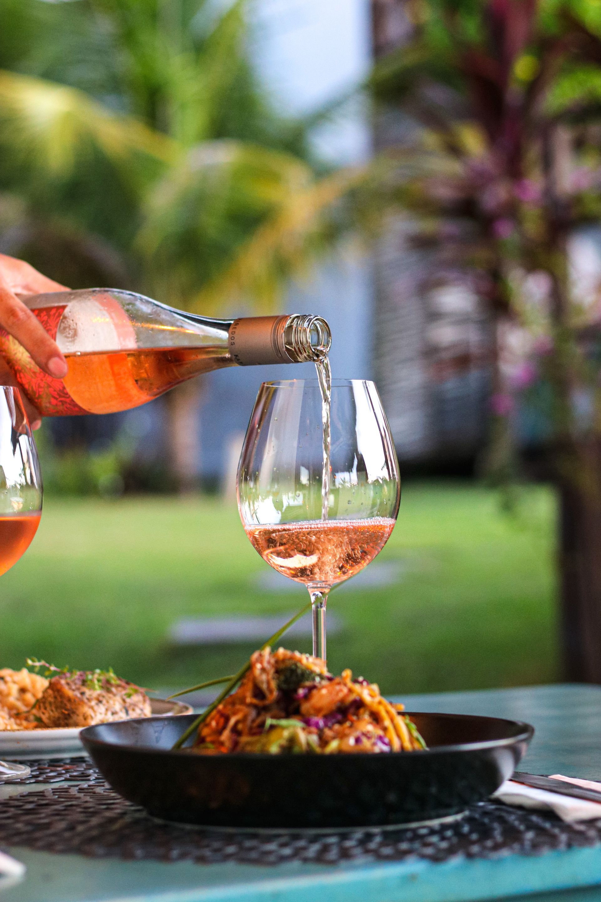 Vinho rosé sendo servido em uma taça sobre uma mesa com uma tigela de comida em um jardim ao ar livre.