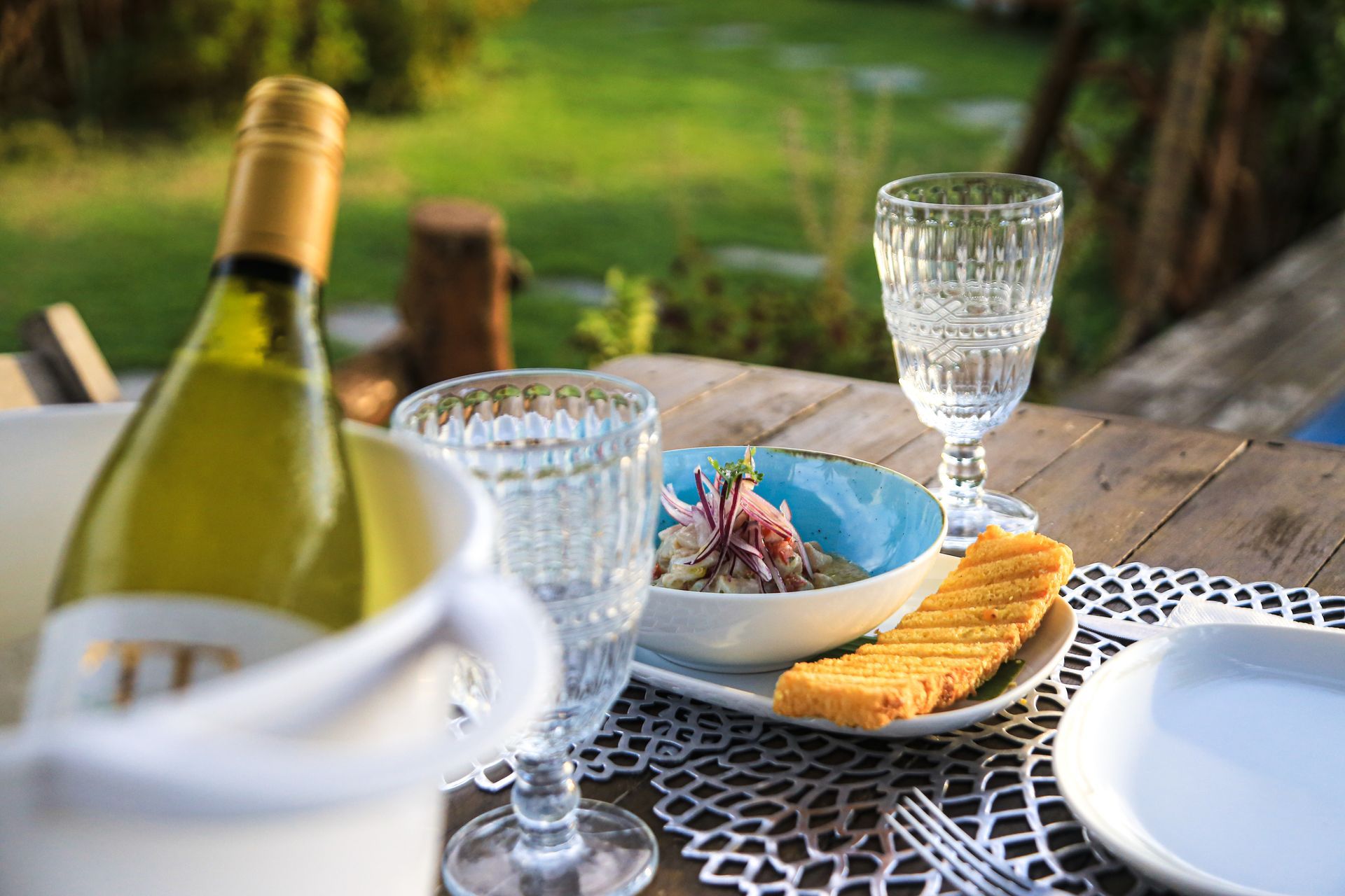 Uma garrafa de vinho branco em um balde de gelo, uma pequena tigela de salada com pão e taças de vinho sobre uma mesa de madeira ao ar livre.