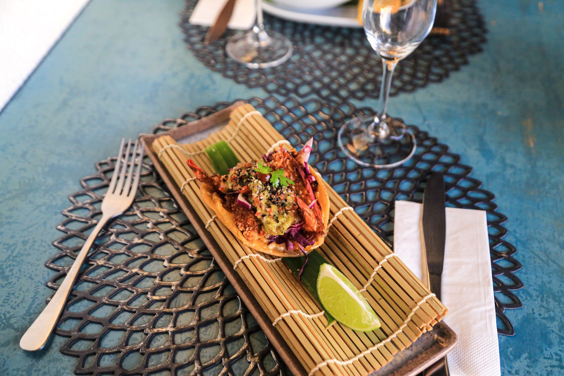 Um taco individual servido sobre uma esteira de bambu com uma fatia de limão, garfo, faca e uma taça de vinho em uma mesa azul texturizada.