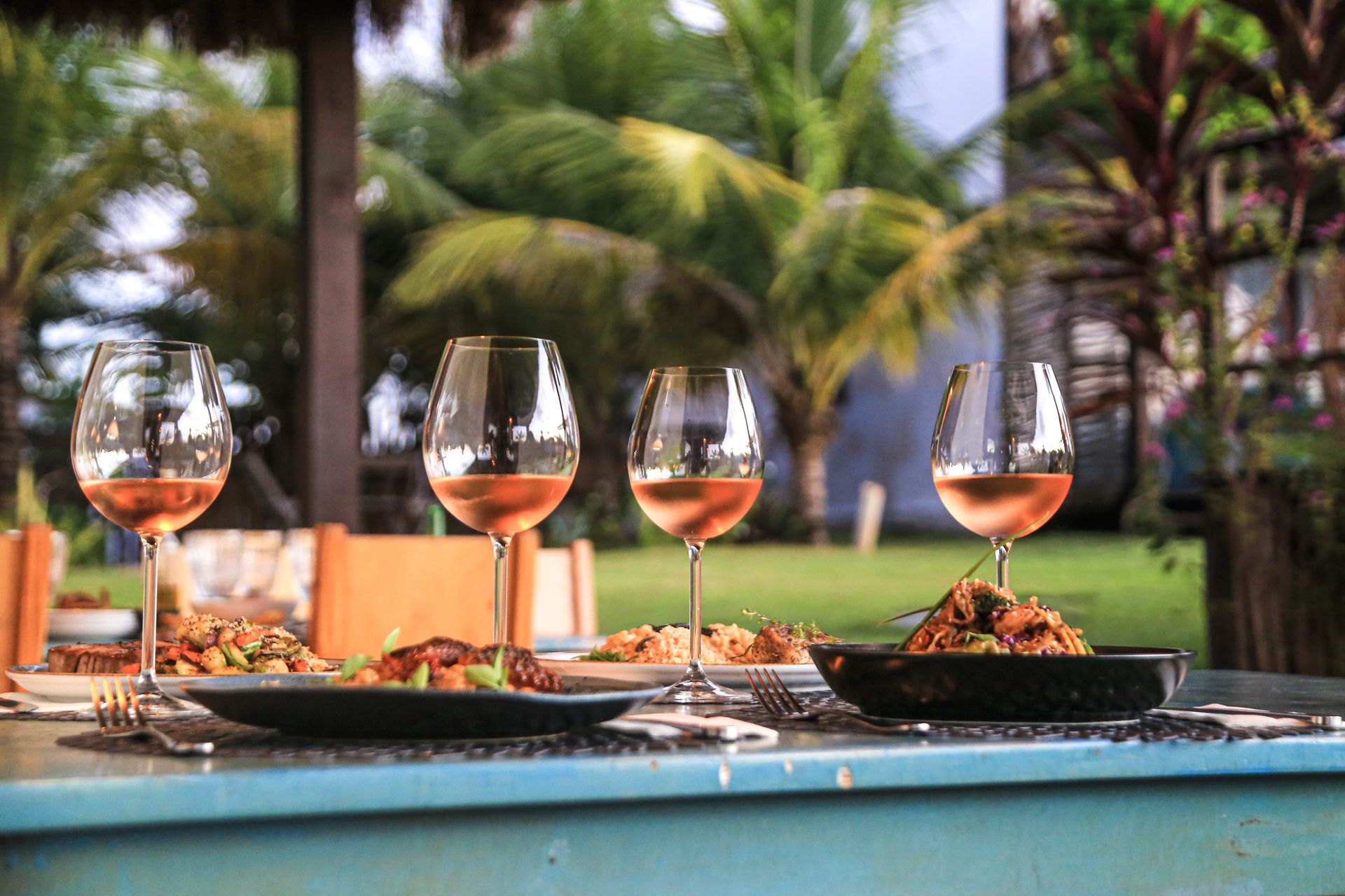 Quatro taças de vinho rosé enfileiradas em uma mesa ao ar livre, com tigelas de comida e palmeiras ao fundo.
