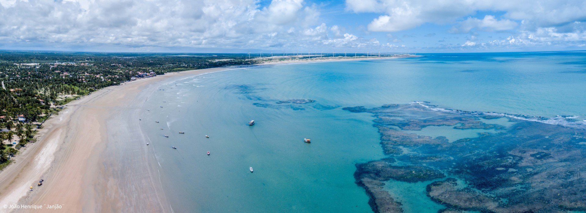 Vista aérea de uma praia costeira com água azul, faixas de areia e recifes rochosos sob um céu claro e nublado.