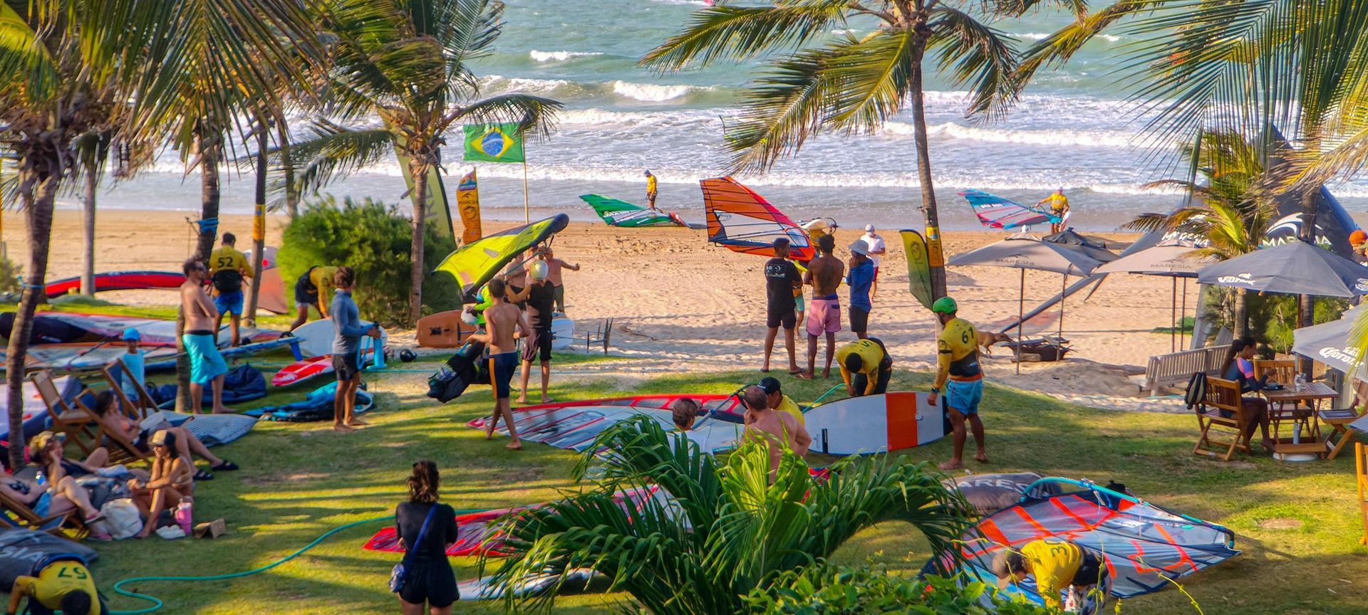 Cena ensolarada de praia com palmeiras, guarda-sóis coloridos e pessoas relaxando perto da costa.
