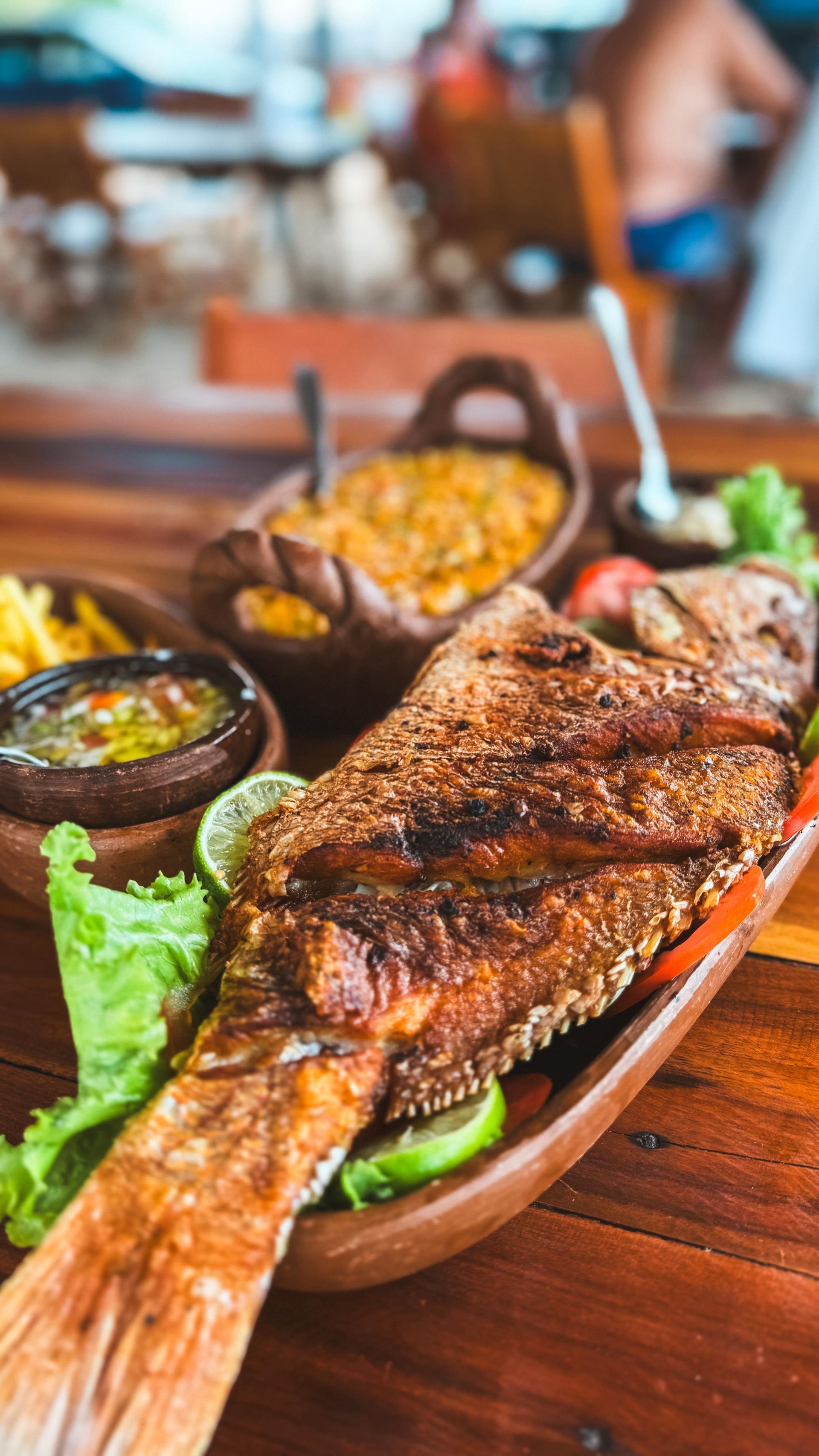 Peixe inteiro grelhado em uma tábua de madeira com acompanhamentos em um ambiente rústico de restaurante.