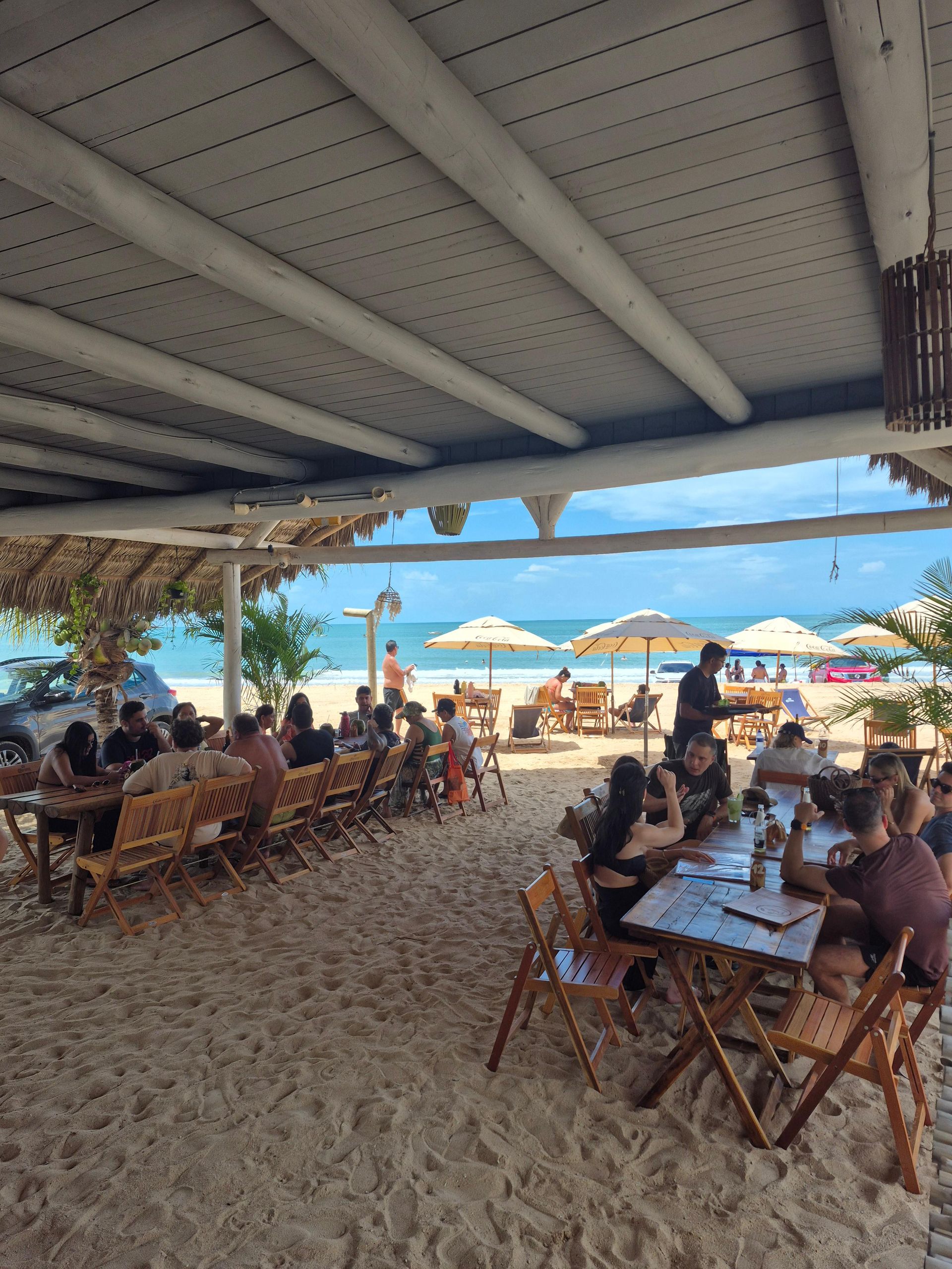 Café à beira-mar com pessoas sentadas sob um telhado sombreado, com vista para o mar e guarda-sóis na areia.