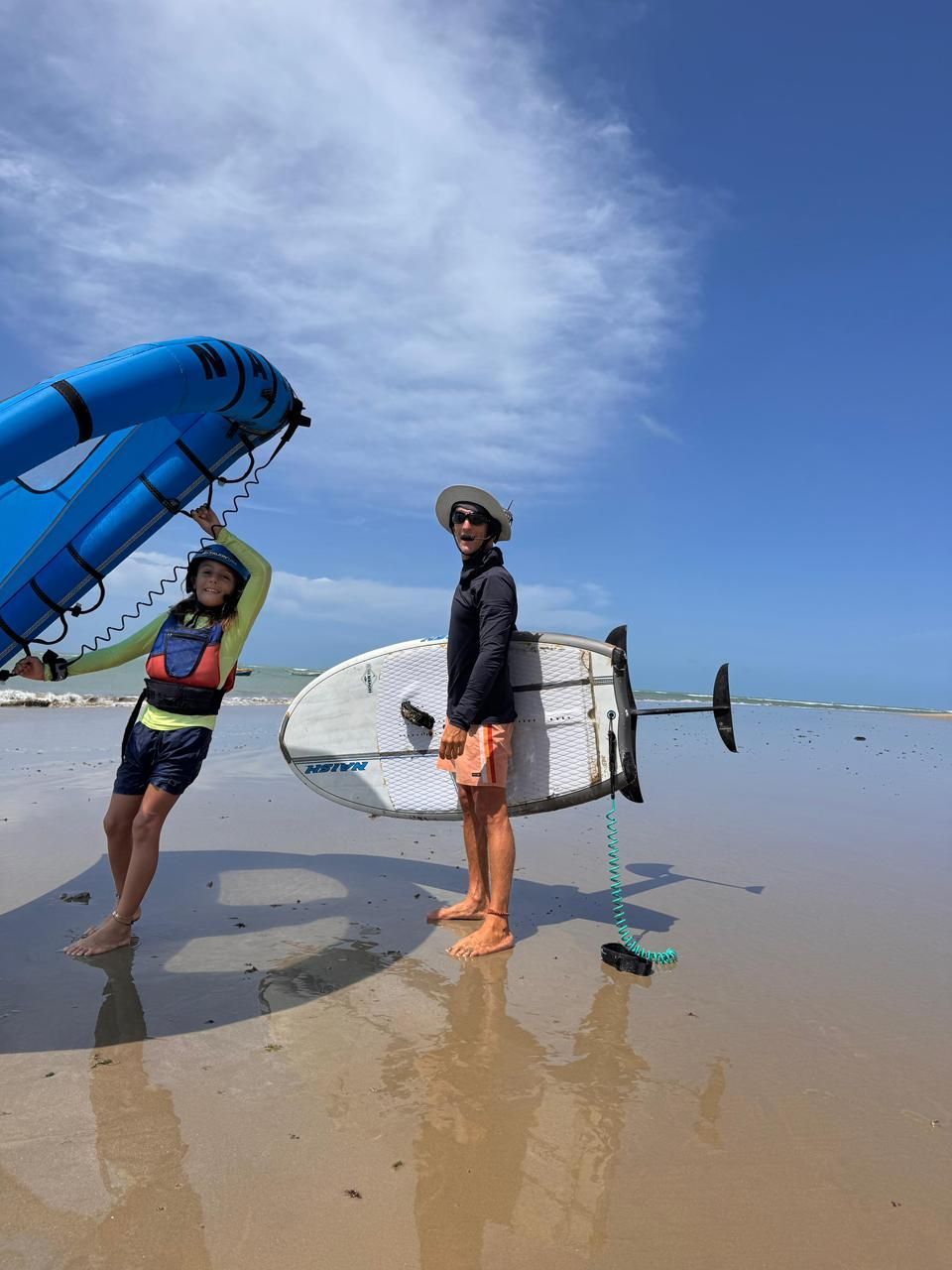 Duas pessoas em uma praia segurando pranchas de surfe ao lado de uma vela azul sob um céu claro.