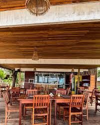 Pátio externo de madeira do restaurante com mesas e cadeiras vazias sob um telhado de palha.