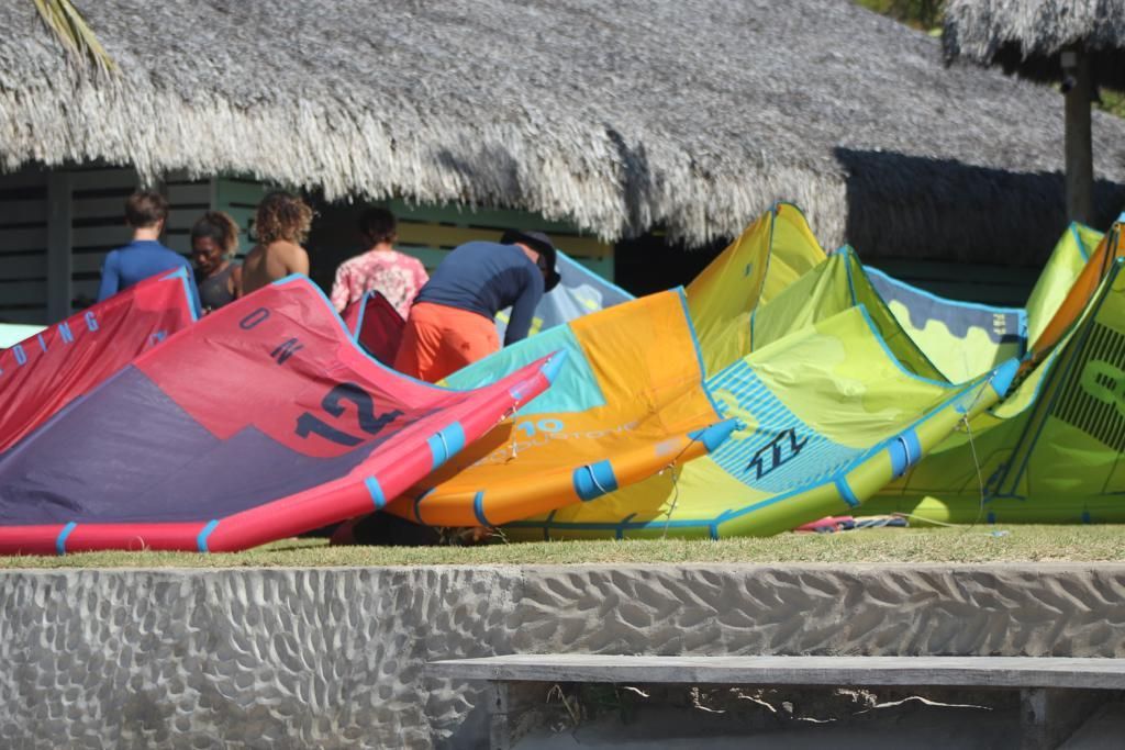 Um grupo de pessoas prepara pipas coloridas de kitesurf em uma área gramada em frente a um edifício com telhado de palha.