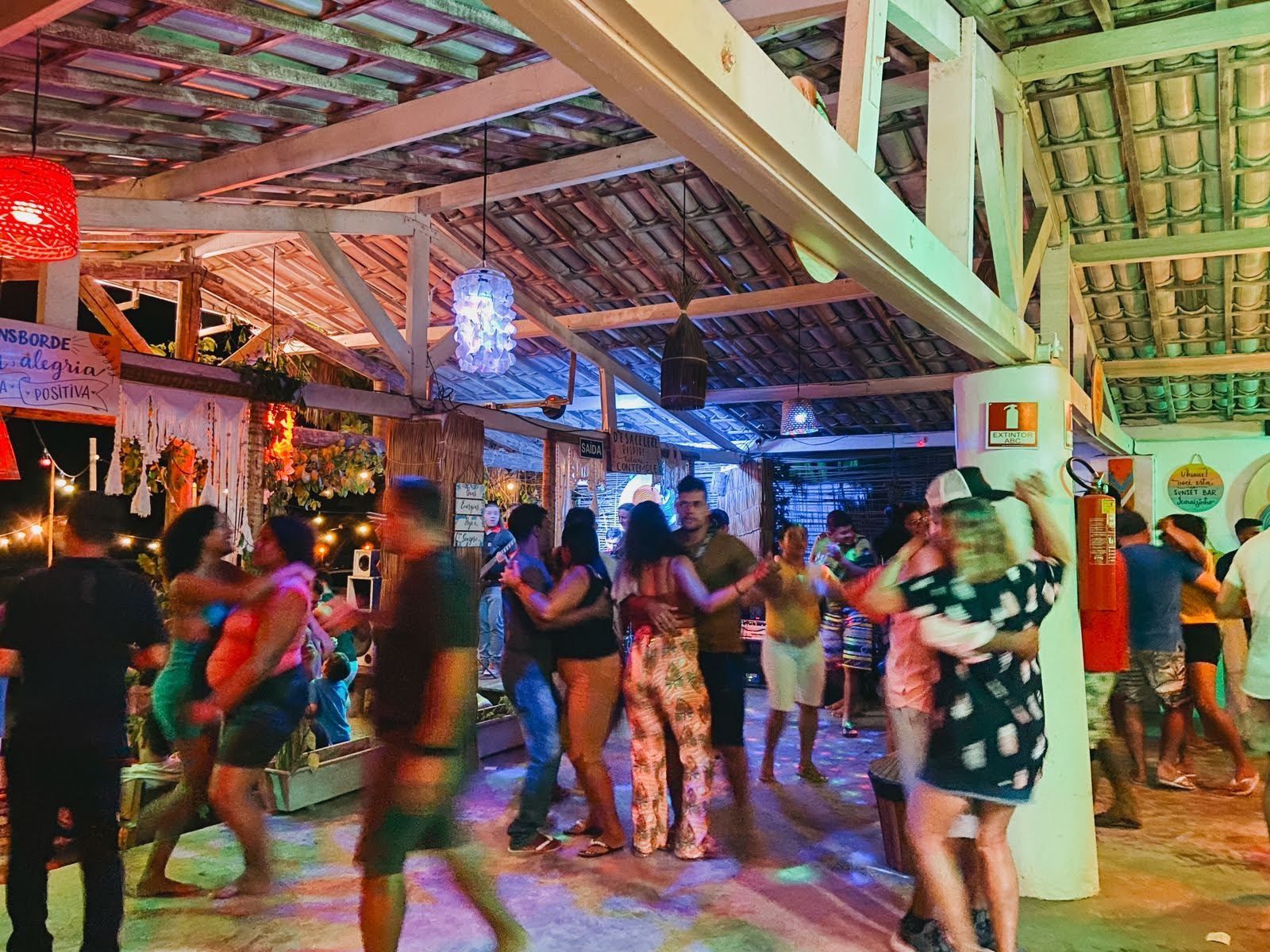 À noite, as pessoas dançam juntas em um restaurante rústico ao ar livre, iluminado por luzes de corda aconchegantes e com teto de palha.