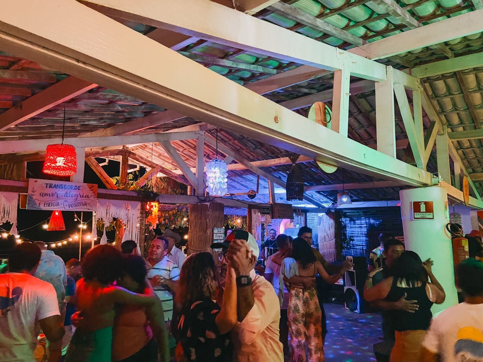 Pessoas dançam em uma festa ao ar livre, à noite, sob um teto de madeira aberto, decorado com luzes festivas vermelhas e brancas.