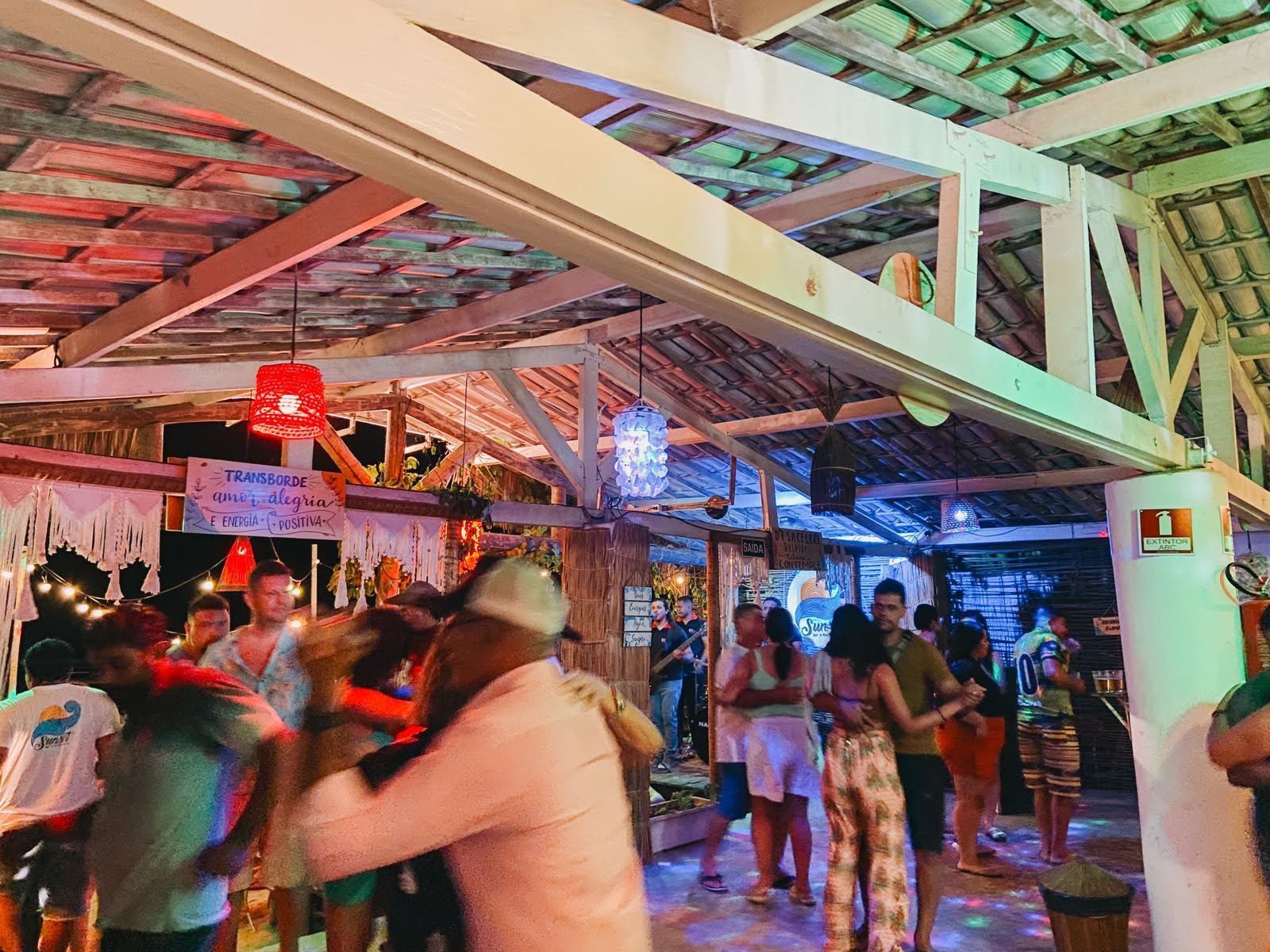 Pessoas dançando em um pavilhão ao ar livre à noite, iluminado por luzes decorativas vermelhas e azuis.
