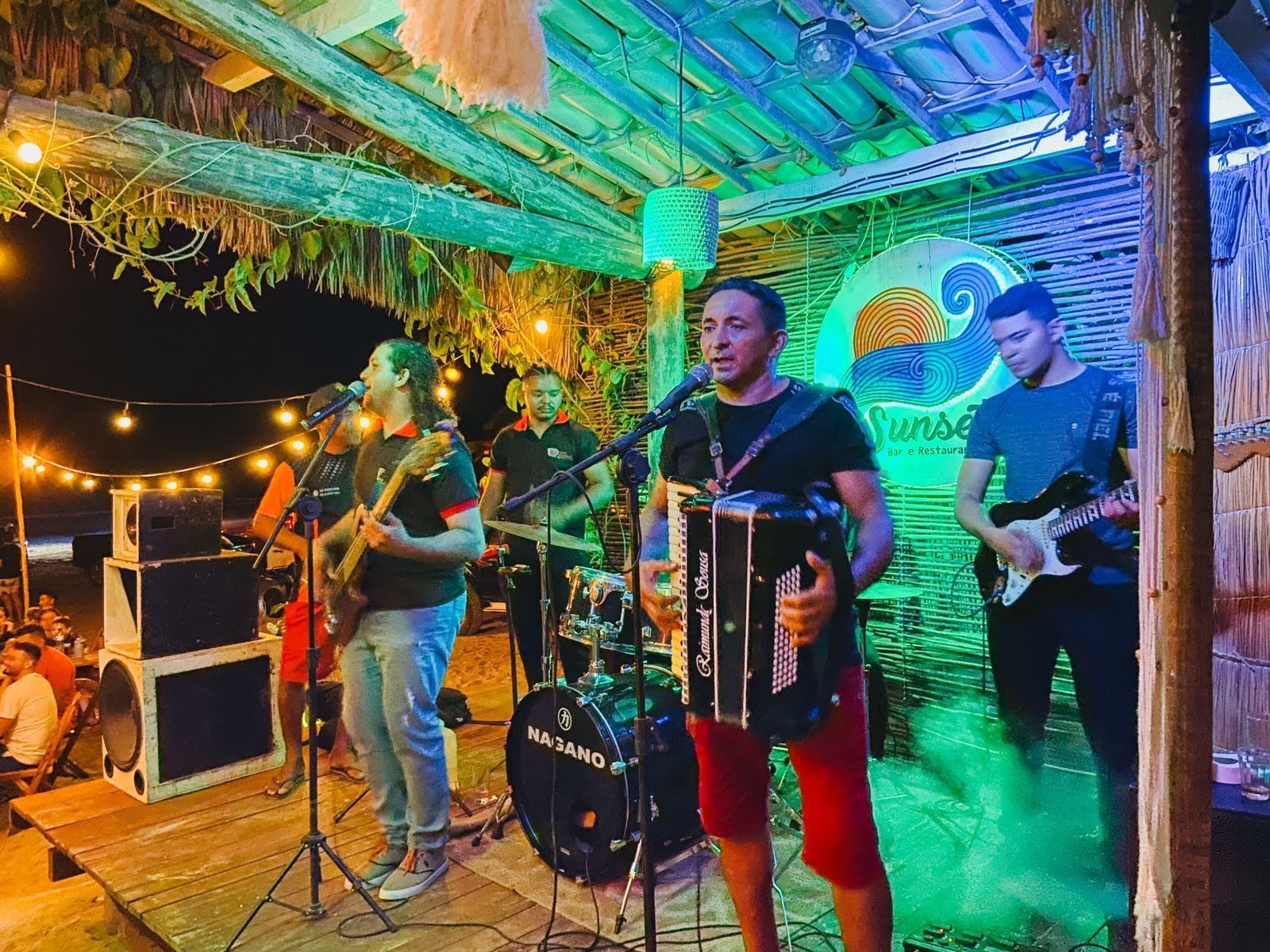 Uma banda se apresenta em um pequeno palco em um local chamado Sunset, composta por um acordeonista, guitarristas e um baterista.