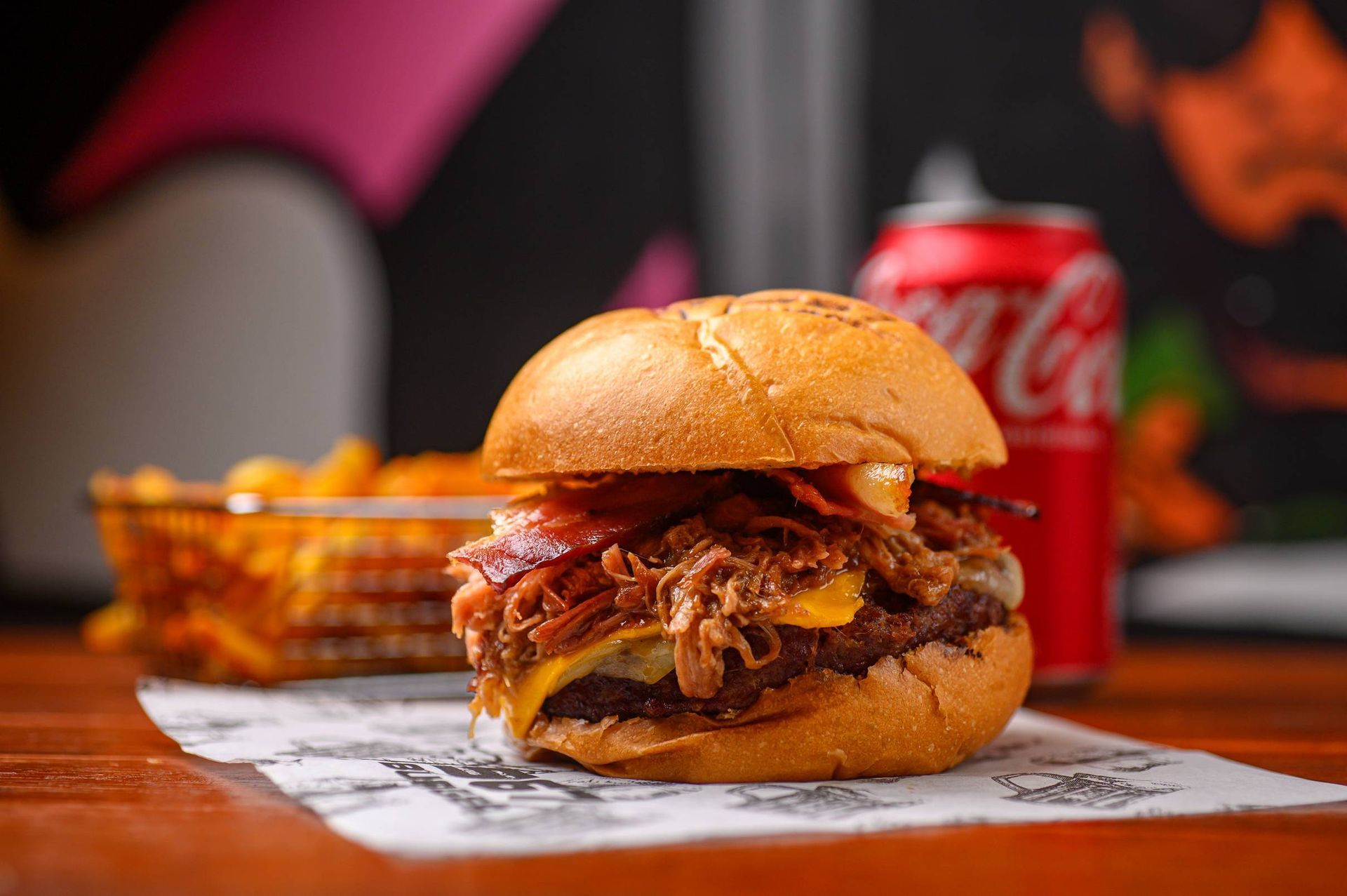 Um hambúrguer gourmet coberto com carne de porco desfiada e bacon está sobre um papel, com uma cesta de batatas fritas e uma lata de refrigerante ao fundo.