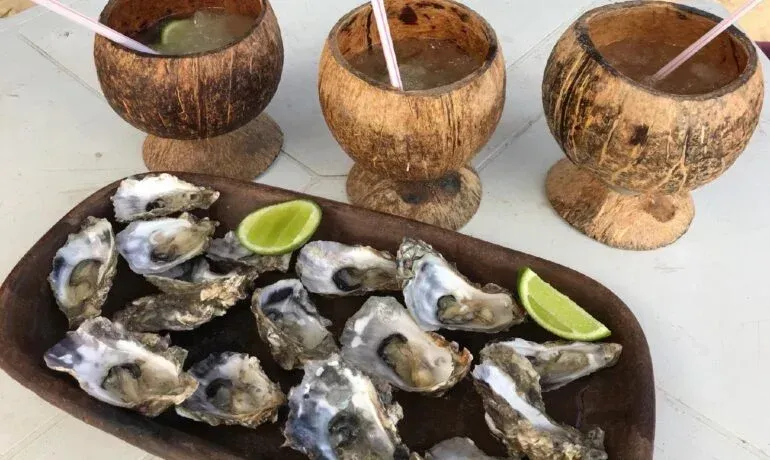 Uma bandeja de madeira com ostras cruas e fatias de limão, acompanhada de três bebidas servidas em cascas de coco sobre uma mesa branca.