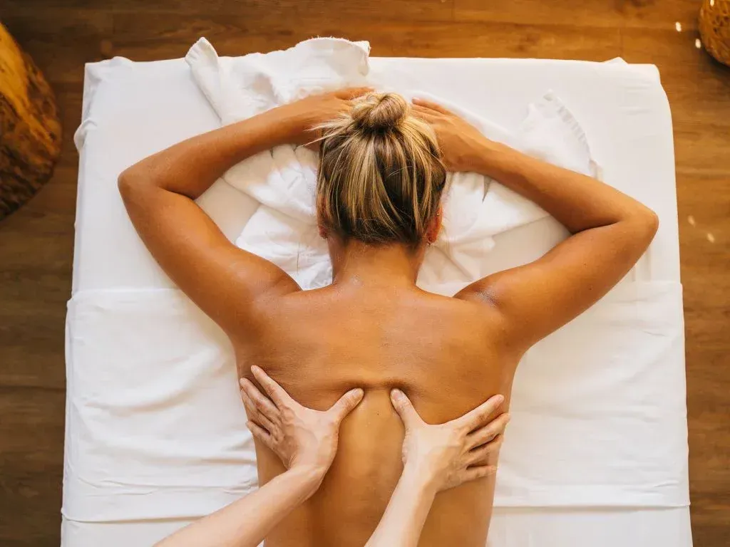 Uma pessoa recebendo massagem enquanto está deitada de bruços em uma maca branca, com as mãos aplicando pressão em suas costas.