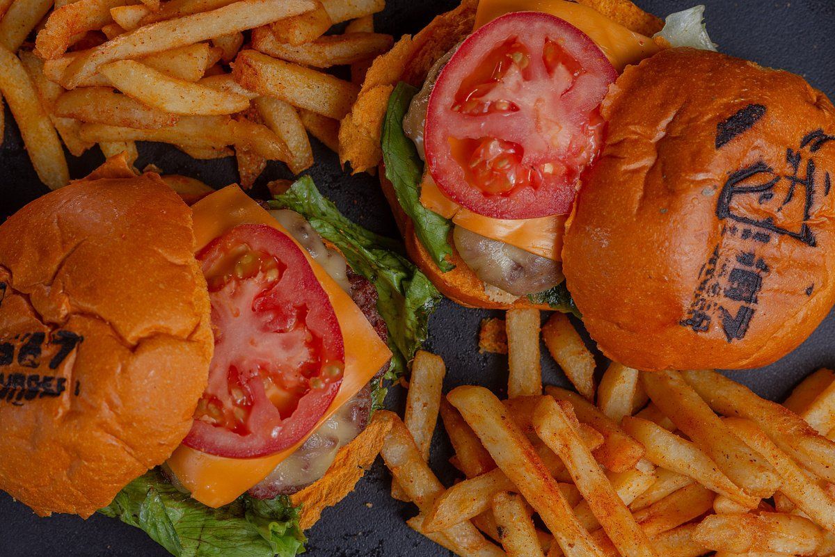 Dois cheeseburgers com rodelas de tomate e alface, servidos com uma porção de batatas fritas temperadas em uma superfície escura.