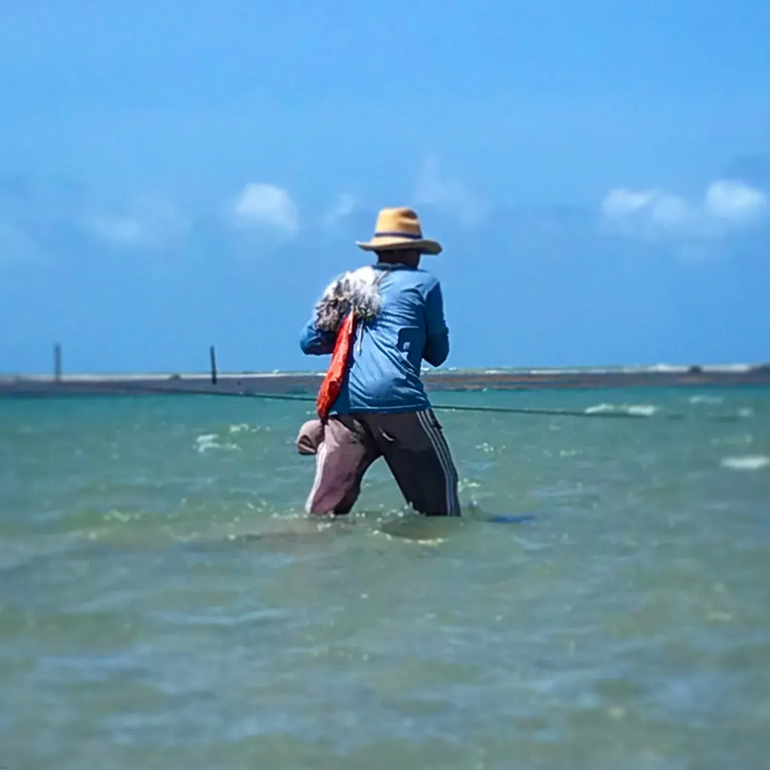 Uma pessoa de chapéu e camisa azul caminhando em águas rasas do oceano, segurando uma rede com cabo laranja.