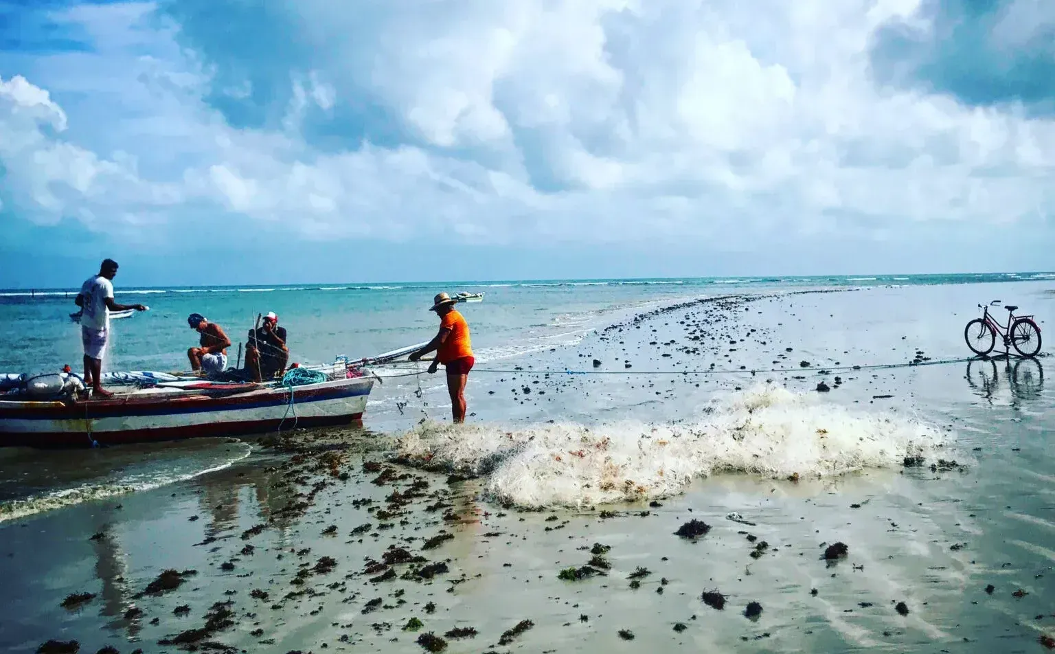 Três pessoas trabalham com um barco de pesca e uma rede em uma praia rasa e arenosa sob um céu nublado, com uma bicicleta por perto.