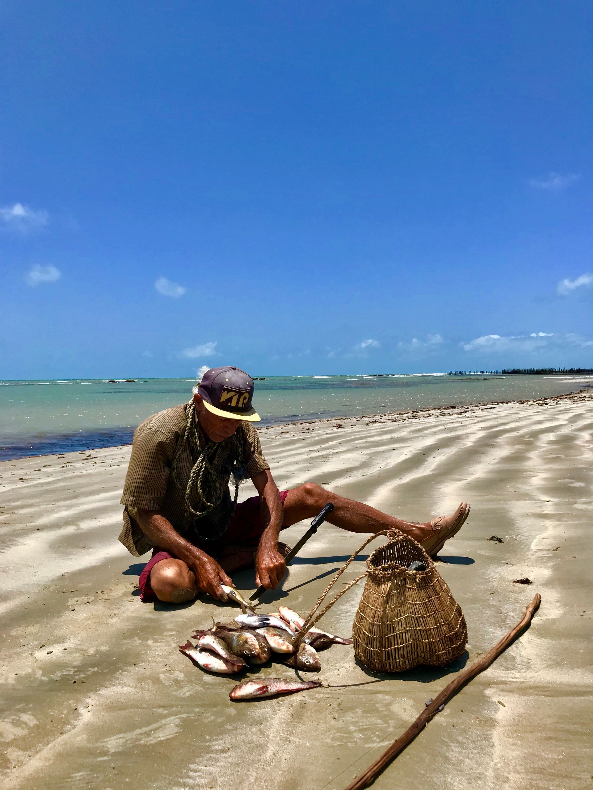 Uma pessoa está sentada na areia da praia, limpando peixes que pescou, ao lado de uma cesta de vime e um longo bastão de madeira.