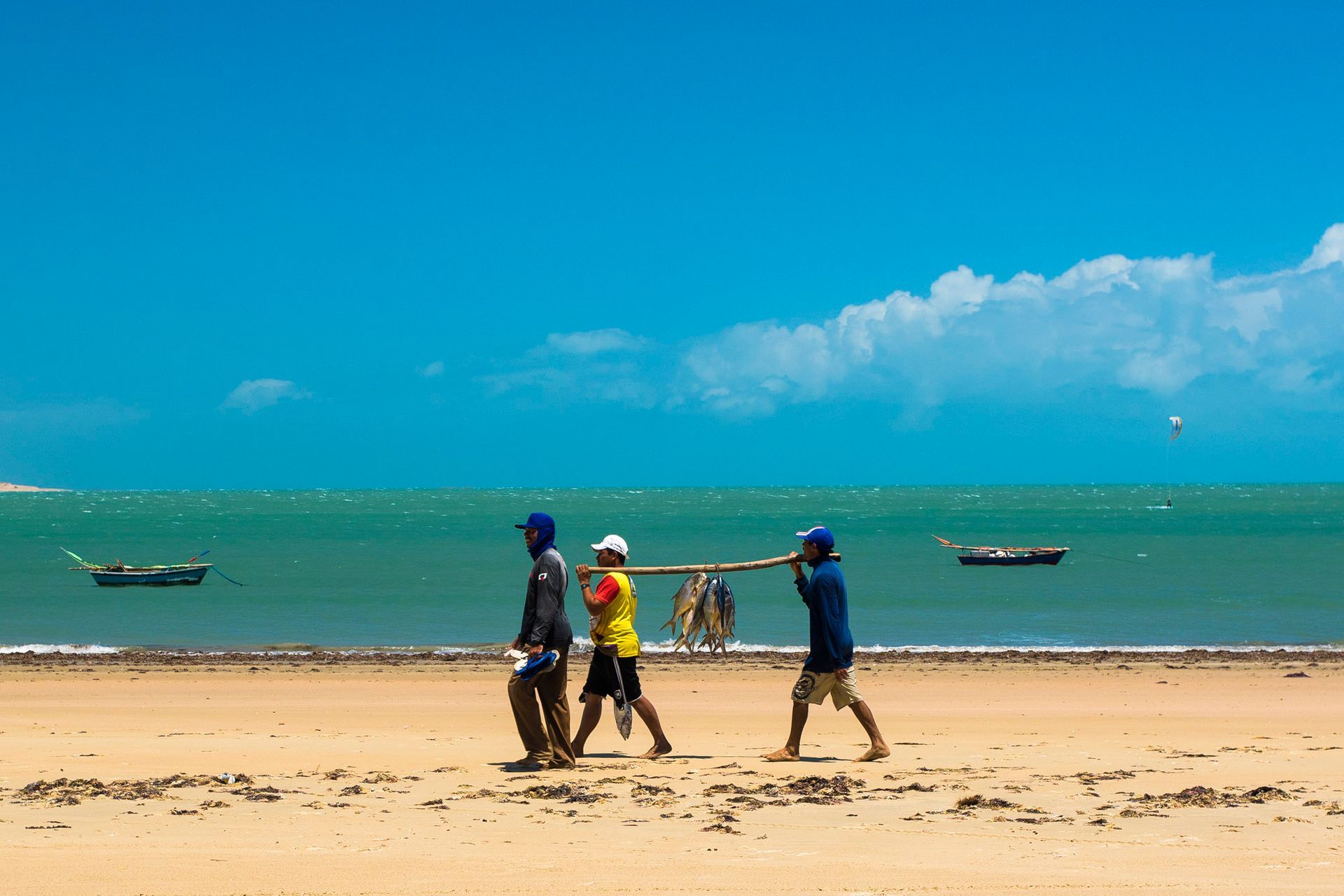 Três pessoas caminham por uma praia de areia carregando uma vara comprida, com pequenos barcos no mar sob um céu azul claro.