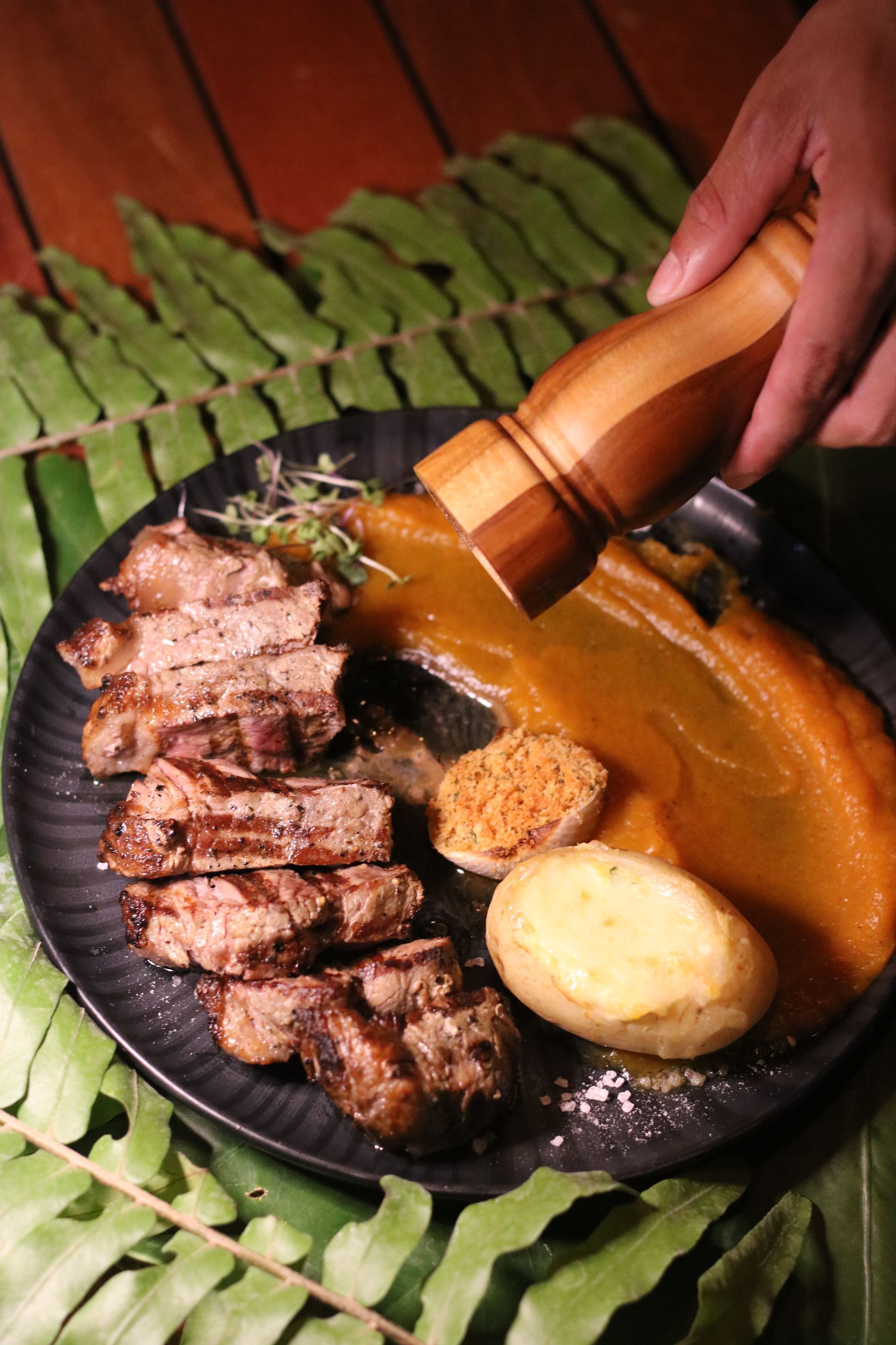 Uma mão mói pimenta sobre um prato de bife grelhado, purê de laranja e uma batata, disposto sobre uma cama de folhas verdes de samambaia.
