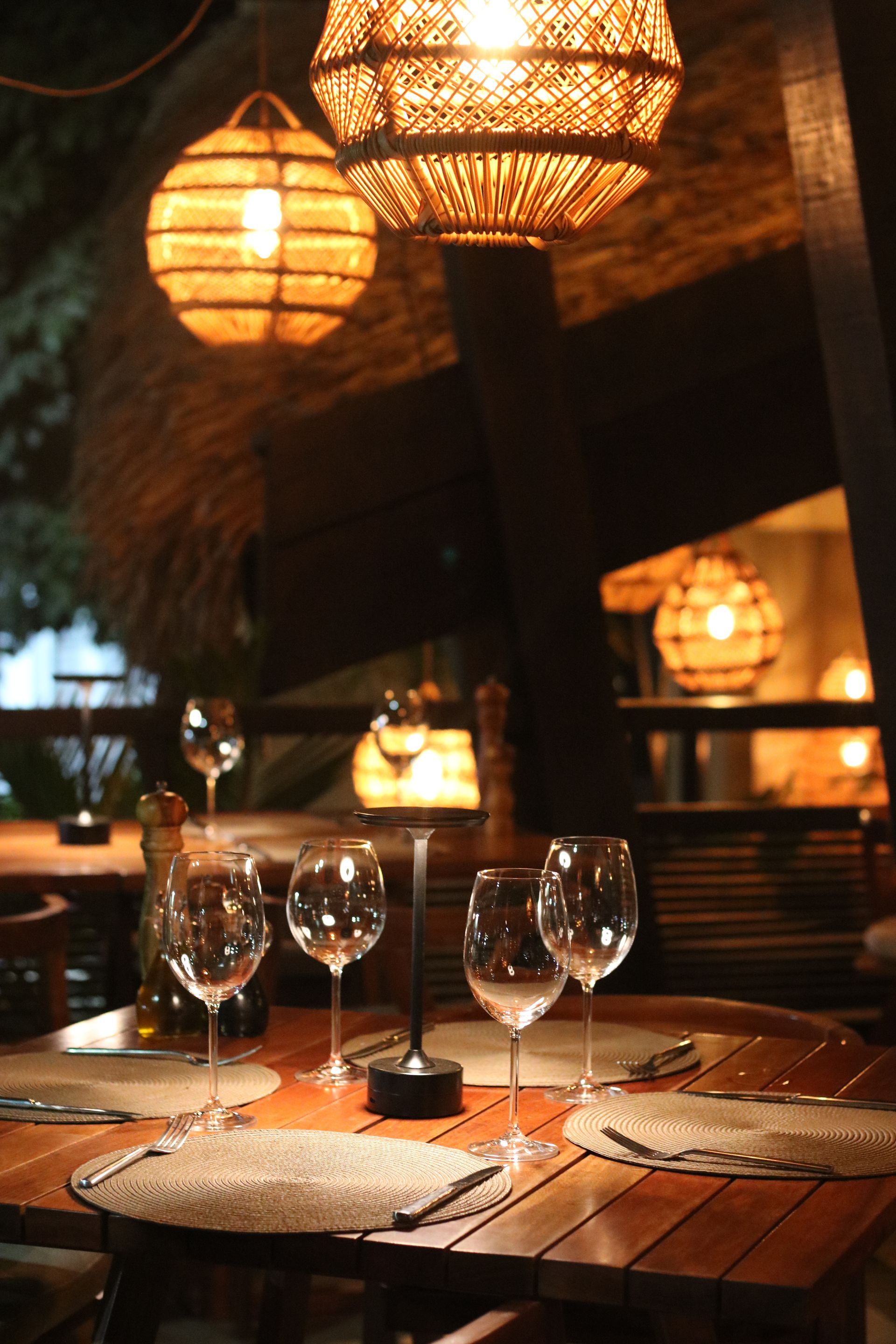 Uma mesa de restaurante com iluminação suave, posta com taças de vinho e jogos americanos de vime, sob luzes de globo suspensas em tons quentes.