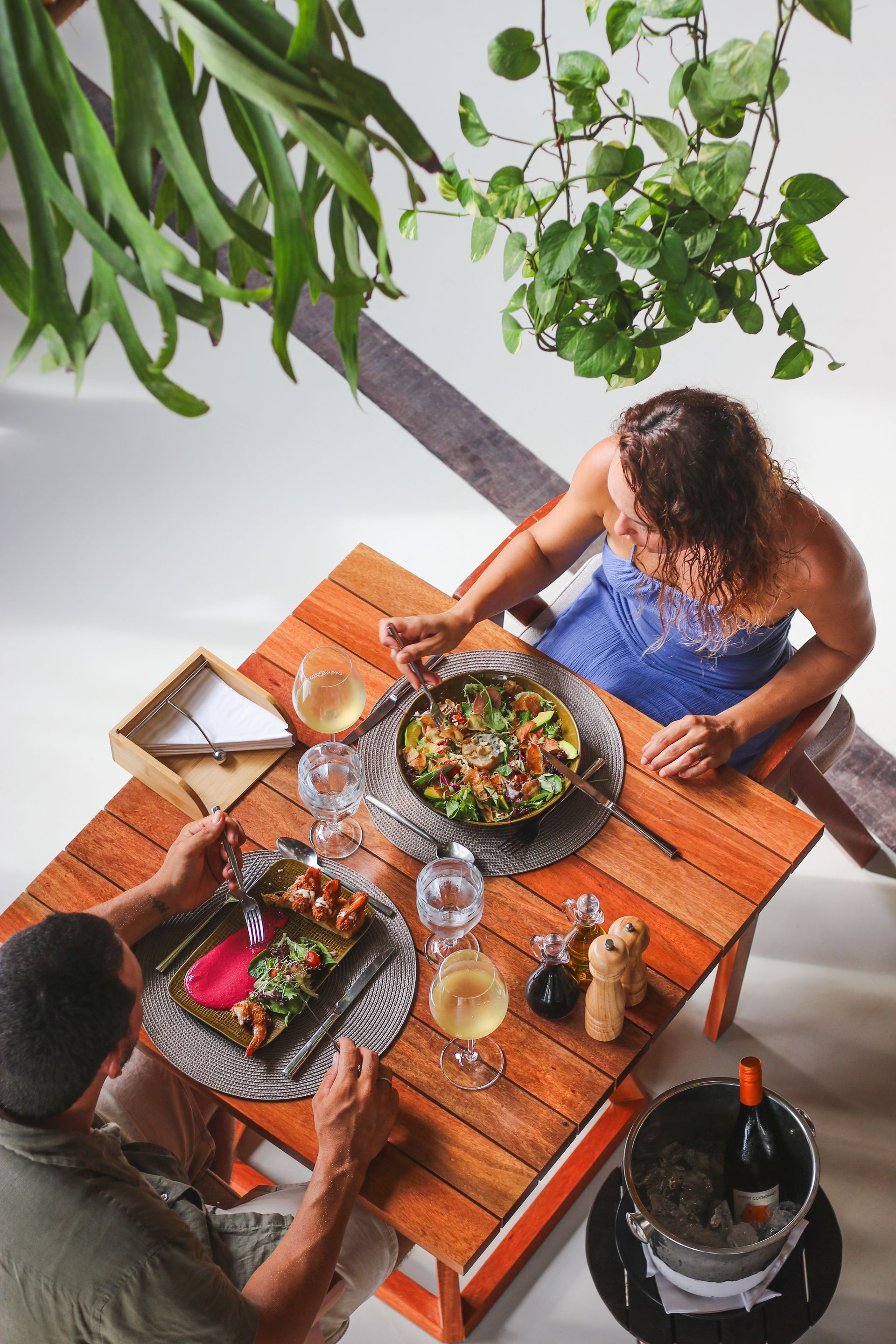 Duas pessoas desfrutam de uma refeição com vinho e um prato principal em uma mesa de madeira ao ar livre, sob plantas suspensas.
