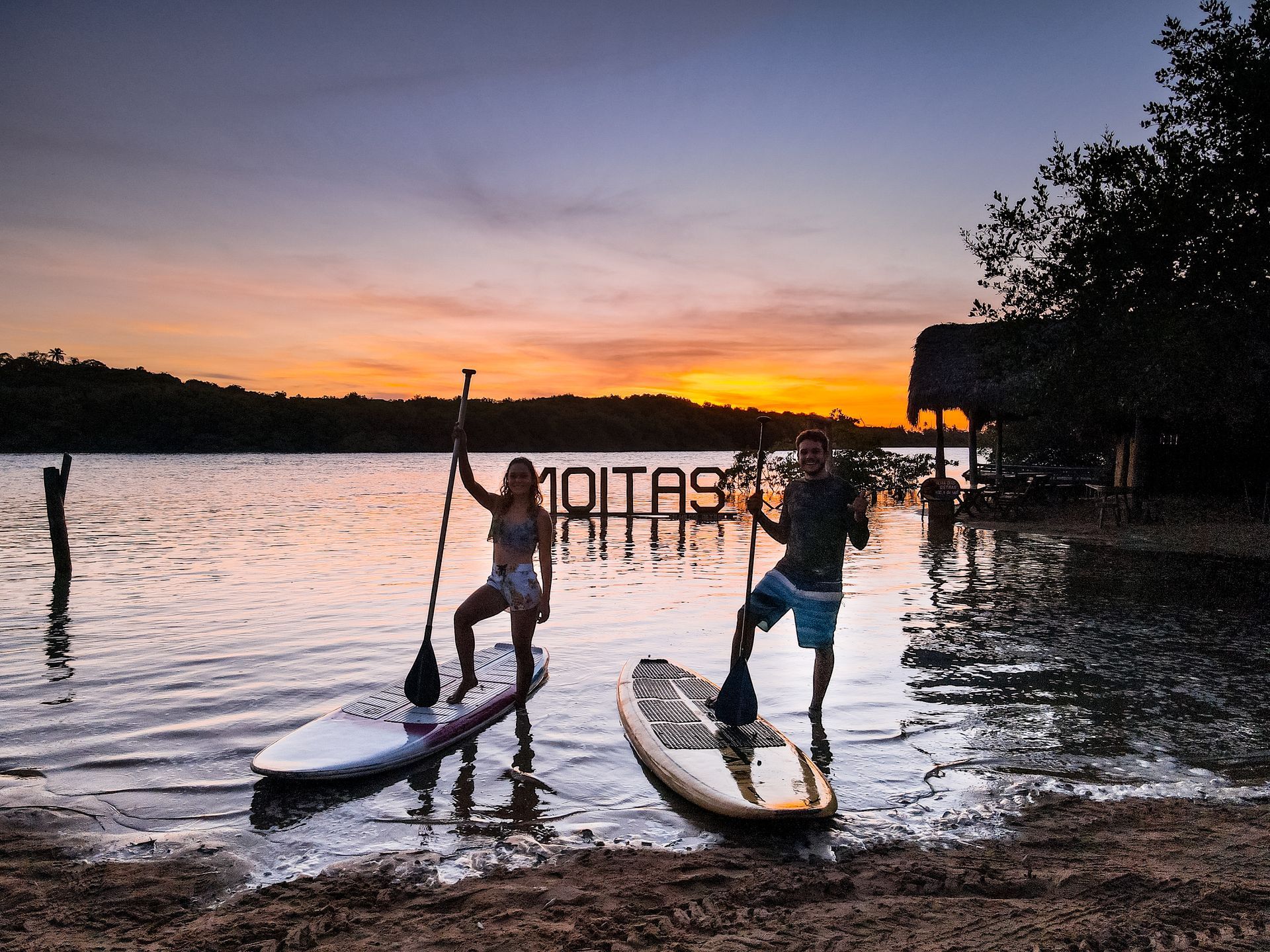 Duas pessoas em pranchas de stand-up paddle estão em águas rasas durante um pôr do sol alaranjado vibrante, com uma placa que diz 