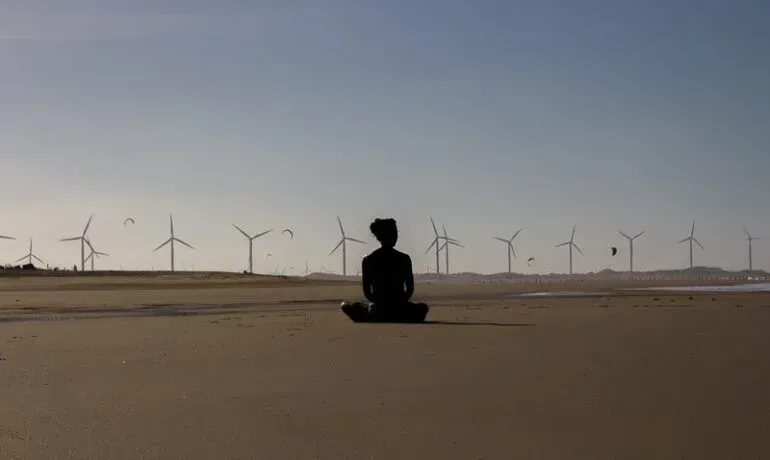 Silhueta de uma pessoa sentada em posição meditativa em uma praia de areia, com inúmeras turbinas eólicas ao fundo.