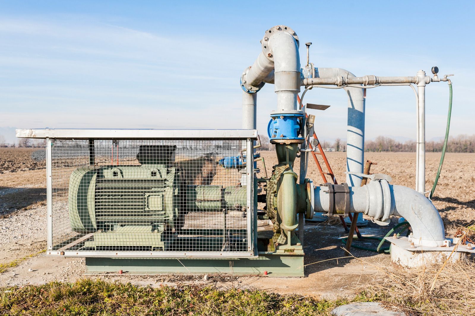 Industrial water pump system on a farm field with metal piping and a protective cage.