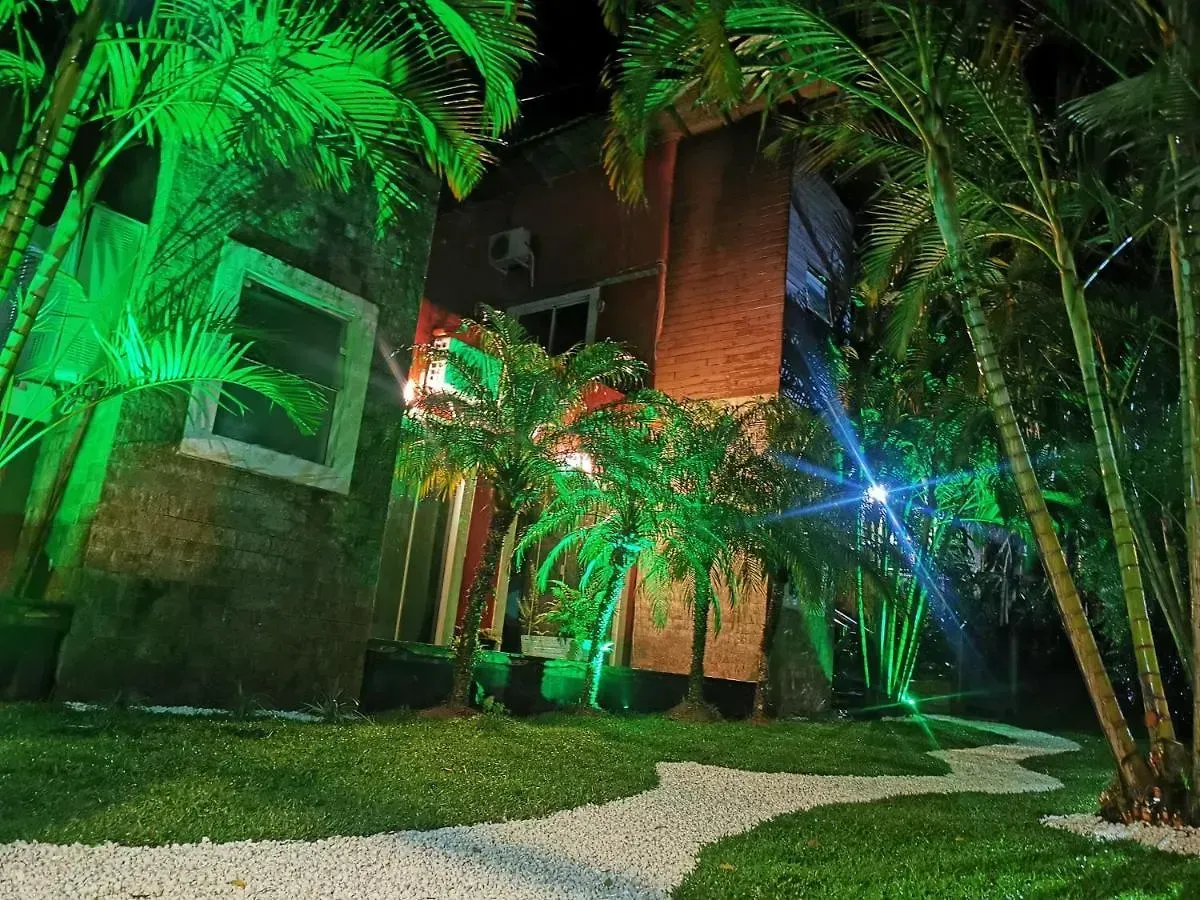 Passarela noturna ao lado de um prédio de tijolos, iluminada por luzes verdes e azuis, com palmeiras acima.