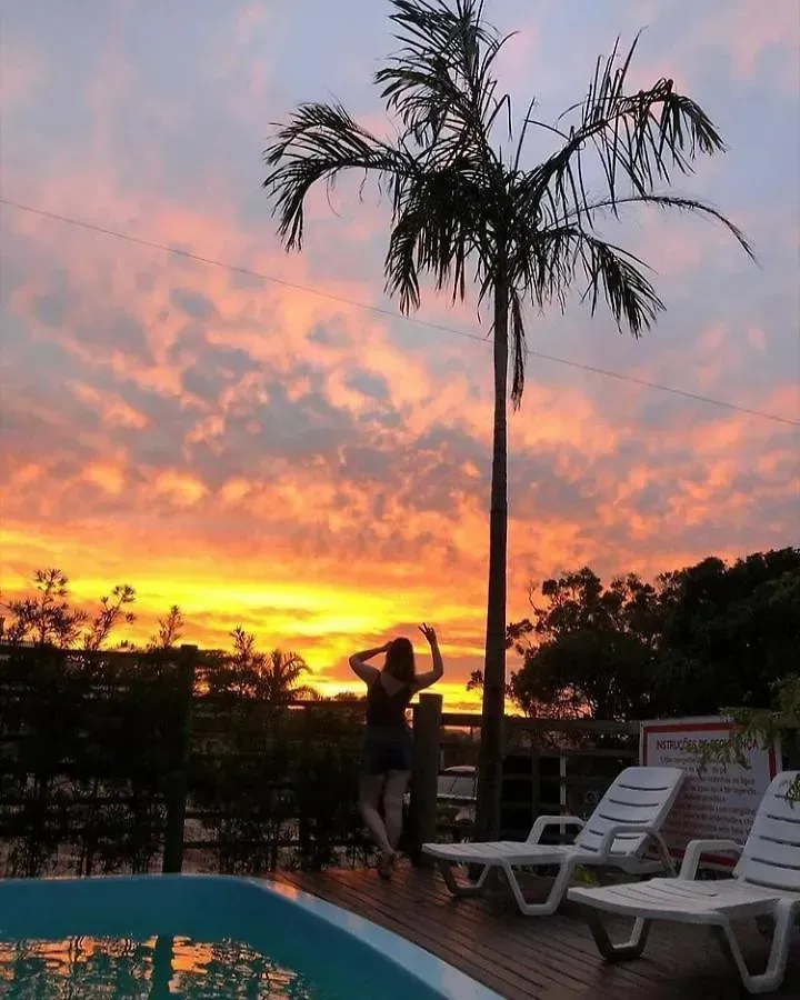 Silhueta de uma pessoa à beira de uma piscina ao pôr do sol, com palmeiras e espreguiçadeiras por perto.