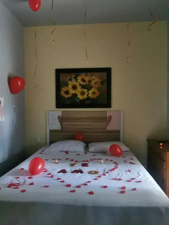 Quarto decorado com uma cama coberta de pétalas de rosa e balões vermelhos, e uma pintura de girassol acima da cabeceira.