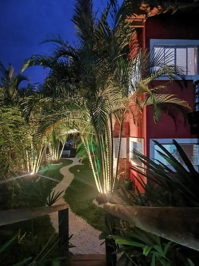 Caminho de jardim ladeado por palmeiras ao entardecer, ao lado de um prédio vermelho com janelas iluminadas e luzes.