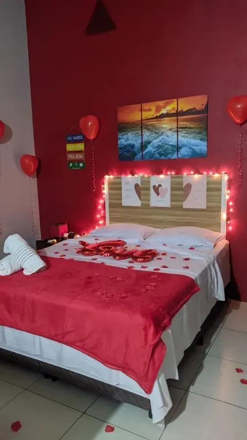 Quarto romântico com paredes vermelhas, balões em formato de coração, pétalas de rosa, luzes de fada e uma cama.