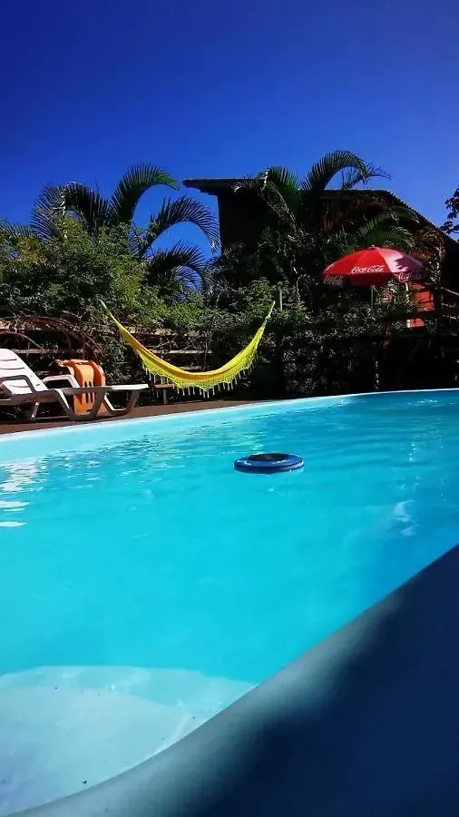 Piscina azul brilhante com uma rede amarela e um guarda-sol vermelho ao lado de palmeiras sob um céu claro.