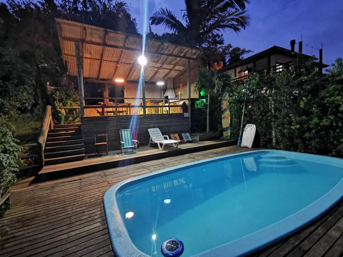 Piscina no quintal ao entardecer, ao lado de um deck de madeira iluminado com espreguiçadeiras, palmeiras e vegetação.
