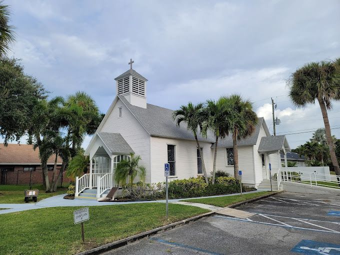 A small white church is surrounded by palm trees and a handicapped parking spot.