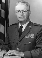 A man in a military uniform and tie is standing in front of a flag.