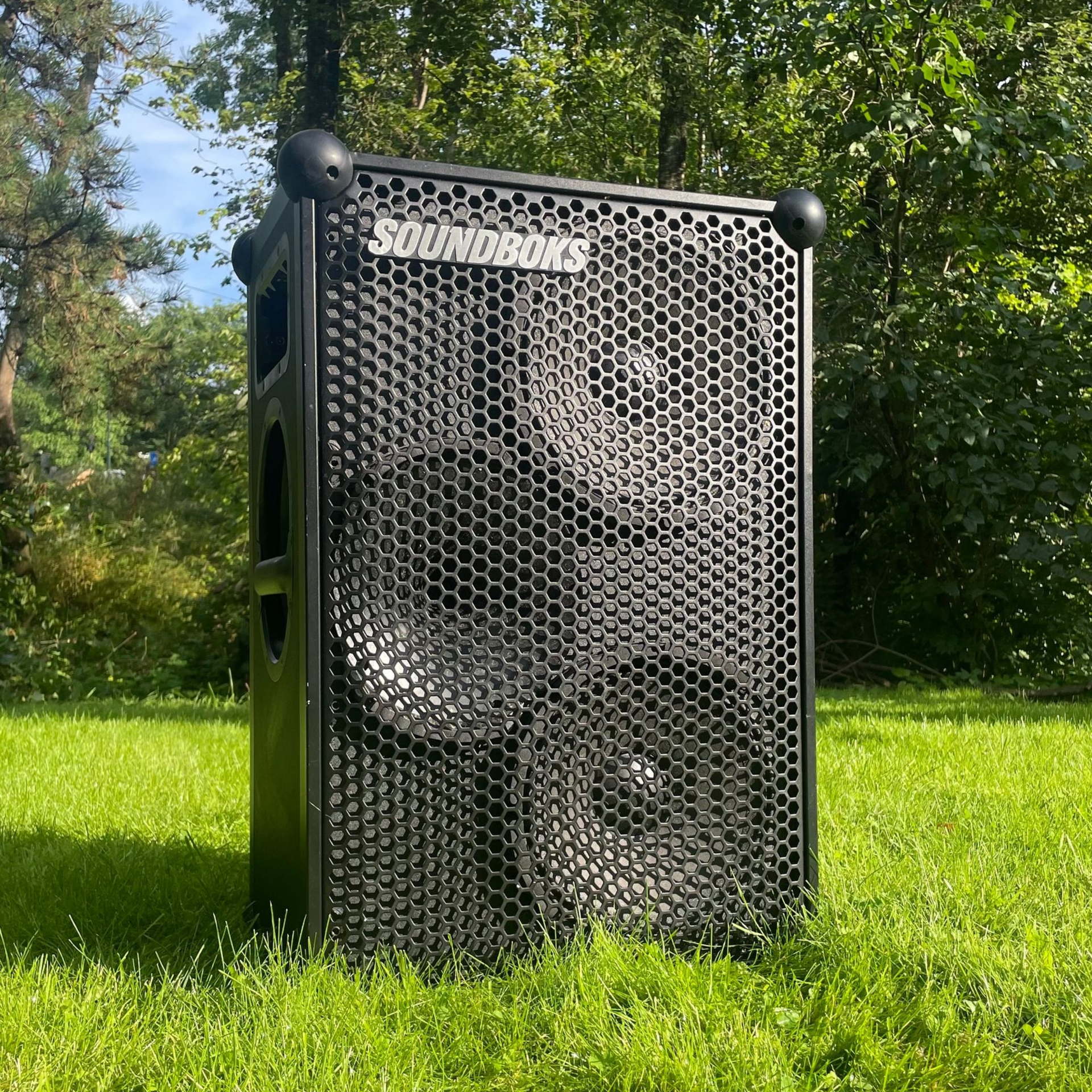 Black speaker on green grass outdoors with a metal grill, logo at the top.