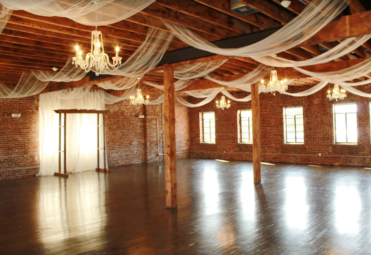 Empty brick-walled hall with polished floor, chandeliers, and sheer fabric draped across the ceiling.