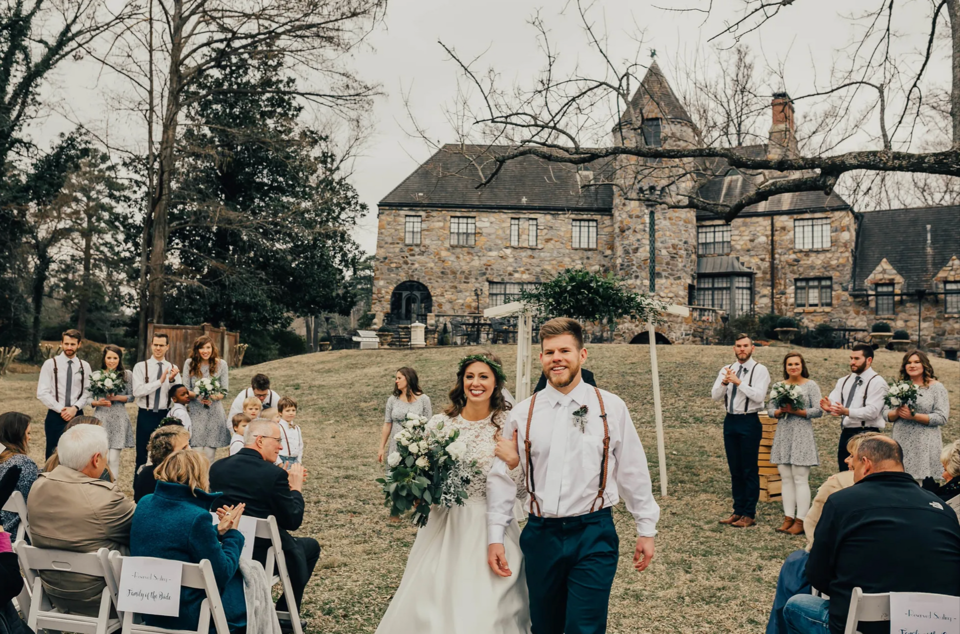 Wedding couple walking down a grassy aisle outdoors, guests seated on both sides before a stone house.