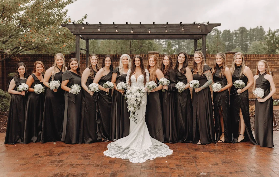Wedding party posing outdoors under a pergola, with the bride in a white gown and bridesmaids in black dresses.