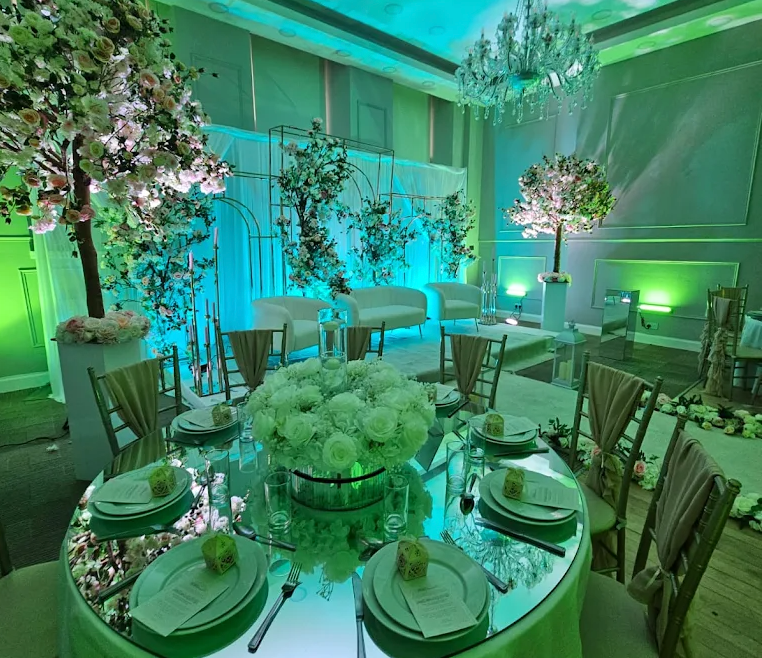 Elegant banquet room with round floral table settings, white chairs, and green-blue ambient lighting