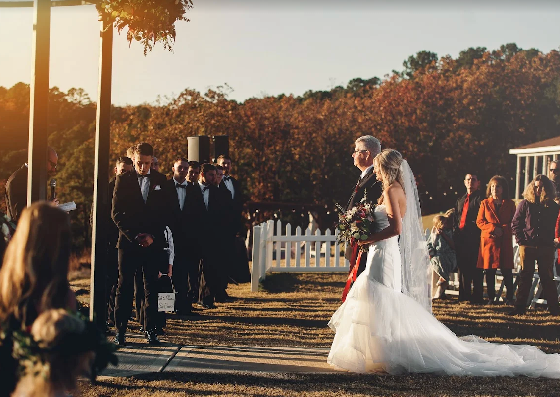 Bride in white gown and veil walking toward guests at an outdoor wedding ceremony at sunset