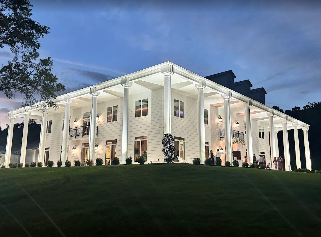 Illuminated white mansion at dusk with tall columns and manicured lawn