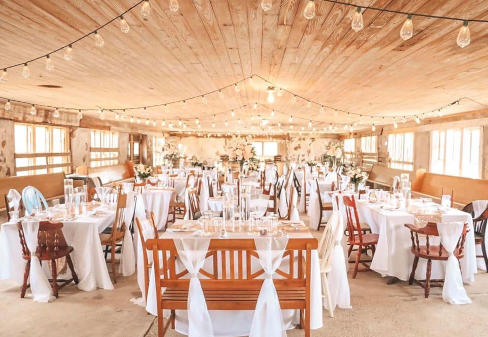 Bright rustic wedding reception hall with round tables, white linens, and wooden chairs under warm string lights